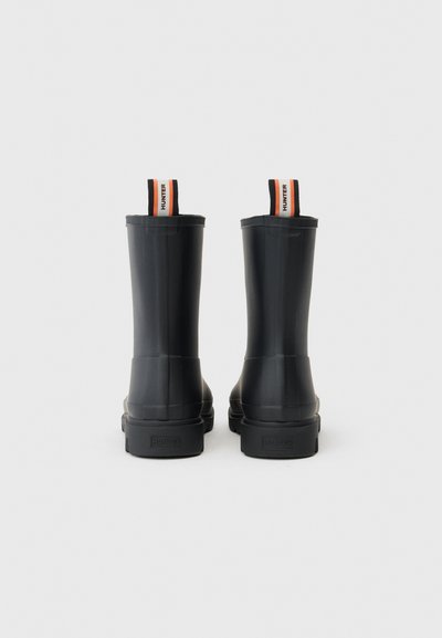 Bottes de cheville en caoutchouc noir avec une texture lisse, dotées de poignées doubles orange et noires, et d'une semelle plate pour plus de stabilité.