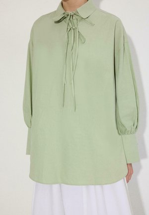 Világos zöld hosszú ujjú blúz, fűzős gallér részlettel, bő fazonú, begyűrt mandzsettákkal, fehér szoknya vagy nadrág fölött viselve.