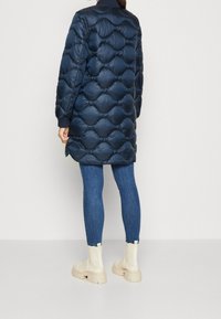 Námořnická modrá péřová bunda s prošívaným designem ve tvaru bubliny, s vysokým límcem a žebrovanými manžetami, nošená přes úzké džíny a krémové kotníkové boty.
