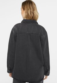Black denim jacket featuring a relaxed fit, long sleeves, a collared neckline, and a coarse texture with a back yoke detail and button cuffs.