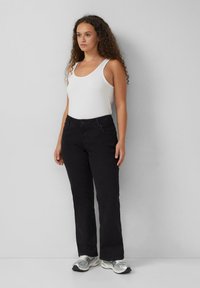 Vrouw met krullend haar, gekleed in een witte tanktop, zwarte jeans en zilveren sneakers, staand tegen een effen lichtgrijze muur.