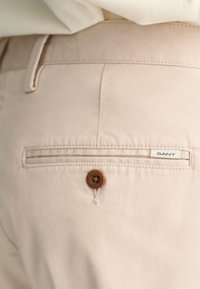 Beige GANT-Hosen mit einer Gesäßtasche, die mit einem braunen Knopf und einem weißen Etikett gesichert ist. Der Stoff ist glatt mit einem dezenten Glanz.