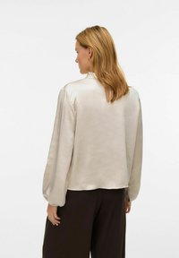 Lichtbeige satijnen blouse met lange mouwen, licht opgeblazen schouders en een keyhole-detail aan de achterkant. Soepele textuur met een ontspannen pasvorm.