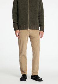 Ελαιόχρωμο φουντωτό πλεκτό φούτερ με ζipper, με ριμπ μανσέτες και στρίφωμα, συνδυασμένο με καφετί κοτλέ παντελόνι και μαύρα δερμάτινα παπούτσια.