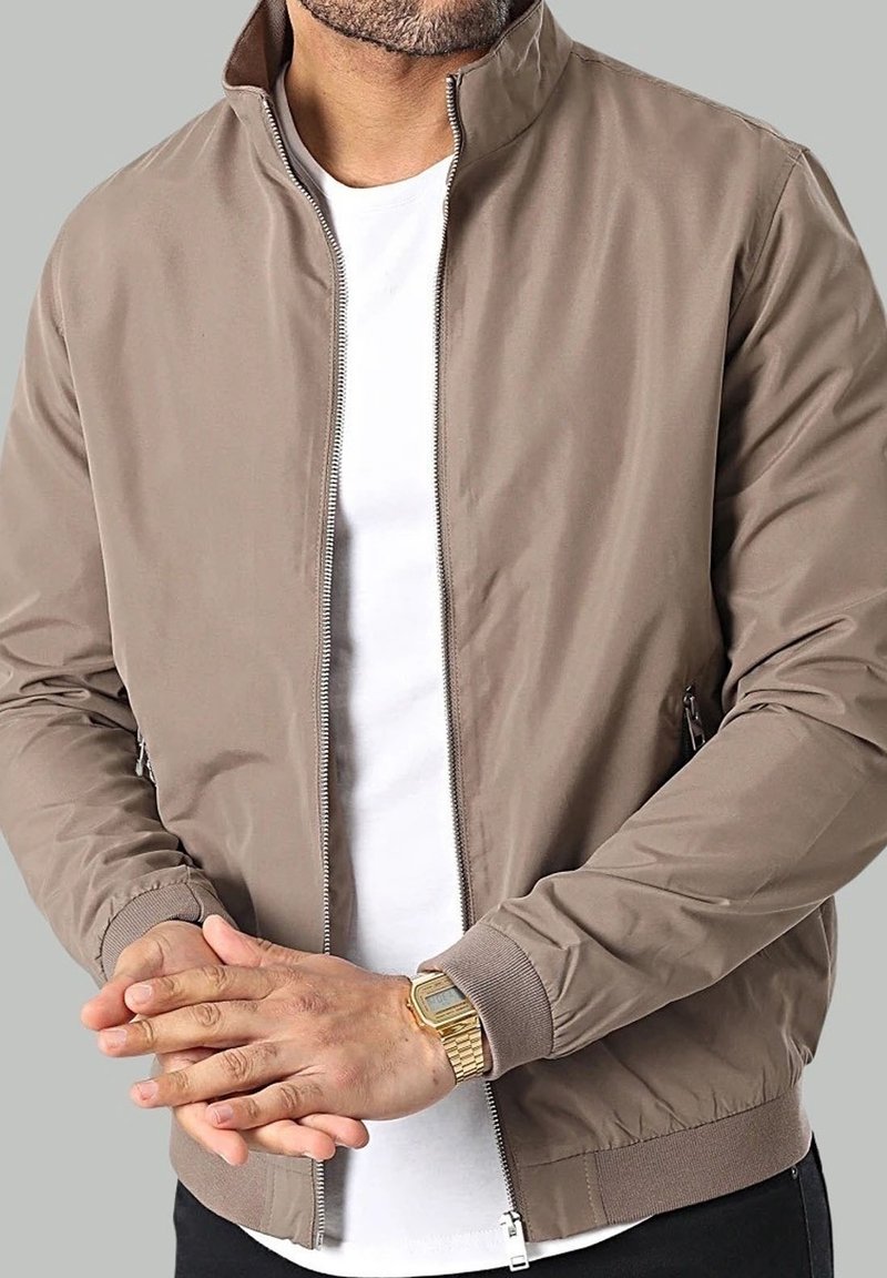 Homme portant une veste beige zippée sur une chemise blanche, avec une montre digitale dorée au poignet, mains jointes, sur fond gris.
