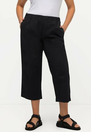 Sorte cropped bukser med elastisk talje, sidelommer og en glat overflade, kombineret med sorte sandaler.