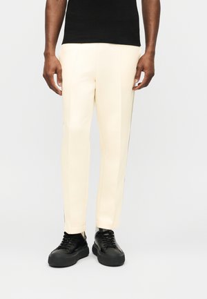 Engedetlen, egyenes szárú, törtfehér nadrágot és vastag talpú fekete tornacipőt viselő személy, egyszerű háttér előtt állva.