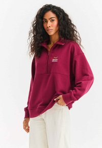Bordowa bluza rozpinana na zamek z długimi rękawami, z przednią kieszenią i wyszywanym złotym logo "Naoko Studio". Miękka faktura tkaniny.