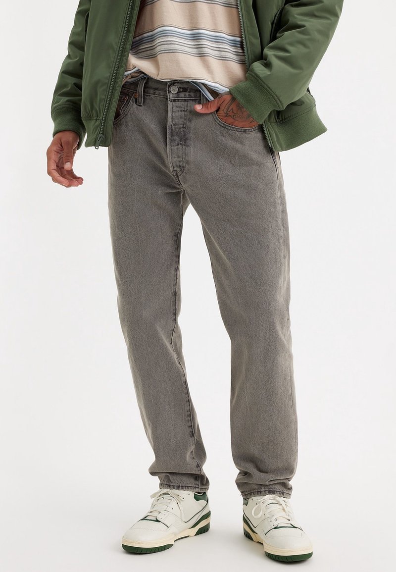 Homme portant un jean gris, des baskets blanches avec des touches vertes, une chemise rayée et une veste verte, debout avec une main dans la poche.