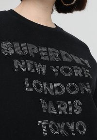 Černá mikina s nápisem "SUPERDRY" a názvy měst zdobenými kamínky. Kolo výstřih a dlouhé rukávy s volným střihem.
