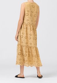 Vestido sin mangas mostaza con recortes florales, diseño con volantes y dobladillo en picos. Llevado con sandalias negras sin talón.