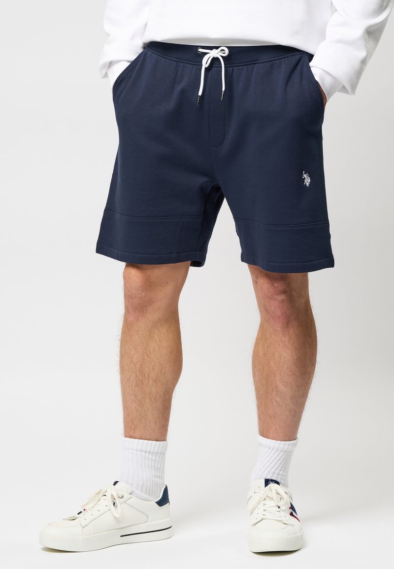 Homme portant un short bleu marine à cordon avec poches latérales, des chaussettes blanches et des baskets blanches sur un fond uni.