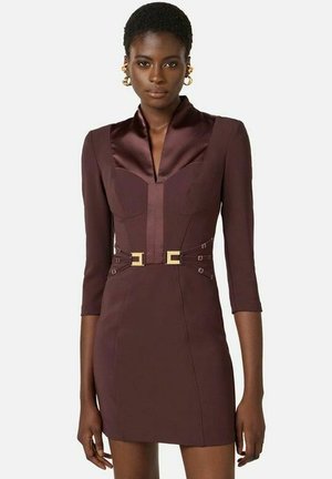 Donna in piedi che indossa un abito aderente marrone scuro con maniche a tre quarti, rever in raso e cintura con fibbia dorata in vita.