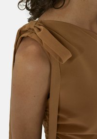 Bruine stof top met een schouderstrikdetail, een gladde textuur en geplooid materiaal aan de zijkant, die een gestructureerd ontwerp benadrukt.