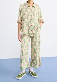 Chemise à manches courtes boutonnée imprimée de motifs floraux en vert et rose, associée à un pantalon large assorti. Chaussures : baskets vertes avec semelles blanches.