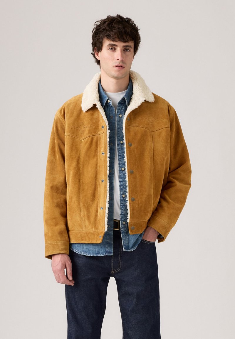 Young man wearing a tan suede jacket with white shearling collar over a blue denim shirt and dark jeans, posing against a plain background.