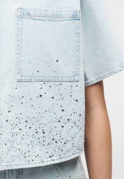 Ανοιχτό μπλε cropped τοπ από denim με τσέπη στο στήθος, διακοσμημένο με διασκορπισμένες μικρές μαύρες και διαφανείς χάντρες κοντά στο τελείωμα.