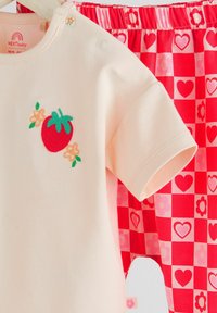 Lys beige bomulds skjorte med broderi af jordbær og blomster, parret med røde ternede shorts prydet med hjerter og blomster i et legende mønster.