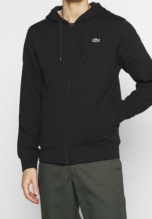 Mand iført sort lynlås hættetrøje med lille grønt krokodille-logo på brystet og olivengrønne bukser, hånd i lommen.