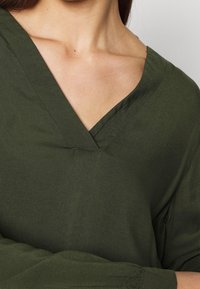 Olivgrünes Shirt mit V-Ausschnitt, aus weichlichem Stoff gefertigt. Es hat lässig geschnittene lange Ärmel mit gerafften Akzenten an den Bündchen.