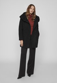 Melna pārdomātas virsjakas ar teksturētu virsmu, lielu apkakli un diviem pogu aizdariem, kas apvienota ar sarkanu turtleneck un melniem plašajiem biksēm.