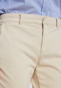 Béžové bavlněné kalhoty s hladkou texturou, s páskem na knoflík a poutky na opasek, částečně zakryté modro-bílou pruhovanou košilí.