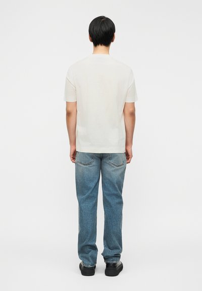 Λευκό κοντομάνικο μπλουζάκι, άνετη γραμμή, συνδυασμένο με ανοιχτό μπλε τζιν παντελόνι, φαίνεται από πίσω με μαύρα παπούτσια. Απλός και μινιμαλιστικός σχεδιασμός.