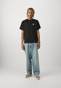 Μαύρο βαμβακερό T-shirt με κοντά μανίκια και λογότυπο μπροστά, συνδυασμένο με ανοιχτό μπλε τζιν παντελόνι σε φαρδιά γραμμή και καφέ αθλητικά παπούτσια. Απλή φόντο.