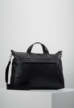 Schwarze Leder-Wochenendtasche mit oberen Griffen und verstellbarem Schulterriemen auf weißem Sockel vor grauem Hintergrund.