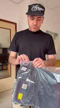 Άνδρας με μαύρο πουκάμισο και καπέλο κρατάει μαύρο ένδυμα σφραγισμένο σε πλαστική συσκευασία μέσα σε κλειστό χώρο.