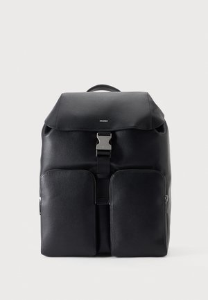 Sac à dos en cuir noir avec un rabat supérieur, une lanière centrale à boucle, une poignée supérieure et deux grandes poches avant, sur fond clair.