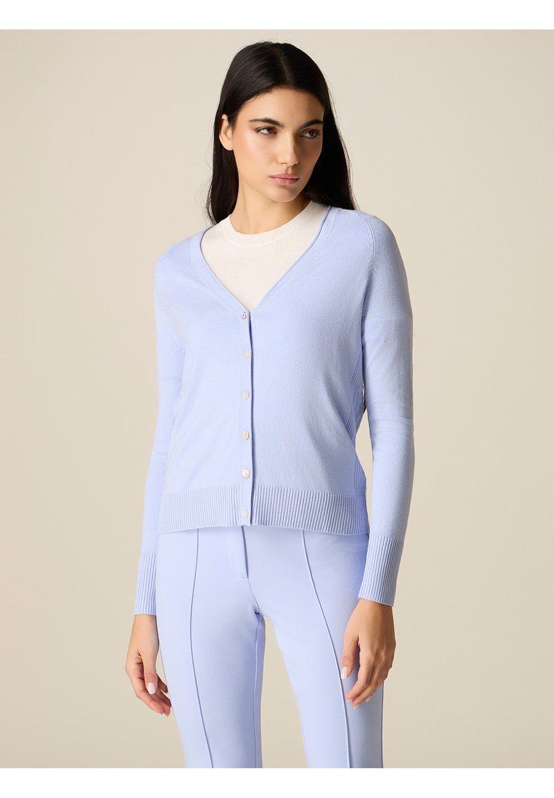 Donna che indossa un cardigan azzurro chiaro abbottonato sopra una maglietta bianca e pantaloni azzurri abbinati, in piedi davanti a uno sfondo beige neutro.