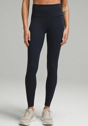 Osoba w czarnych legginsach z wysokim stanem i beżowych butach sportowych, stojąca na jednolitym szarym tle.