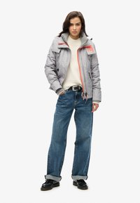 Grå polstret jakke med røde lynlåse, båret over en cremefarvet sweater og parret med brede blå jeans og sorte sko.