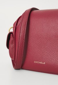 Burgundy leather crossbody bag with pebbled texture. Features a gold-tone logo and strap, rounded edges, and a compact rectangular shape.