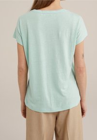 Kortärmad mintgrön t-shirt i mjukt tyg; avslappnad passform med rund halsringning och lätt böjd nederkant, i kombination med beiga byxor.