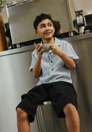 Poiss triibulises särgis ja mustades lühikestes pükstes, istub pingil, hoiab tassi, naeratab moodsas köögis või kohvikus.