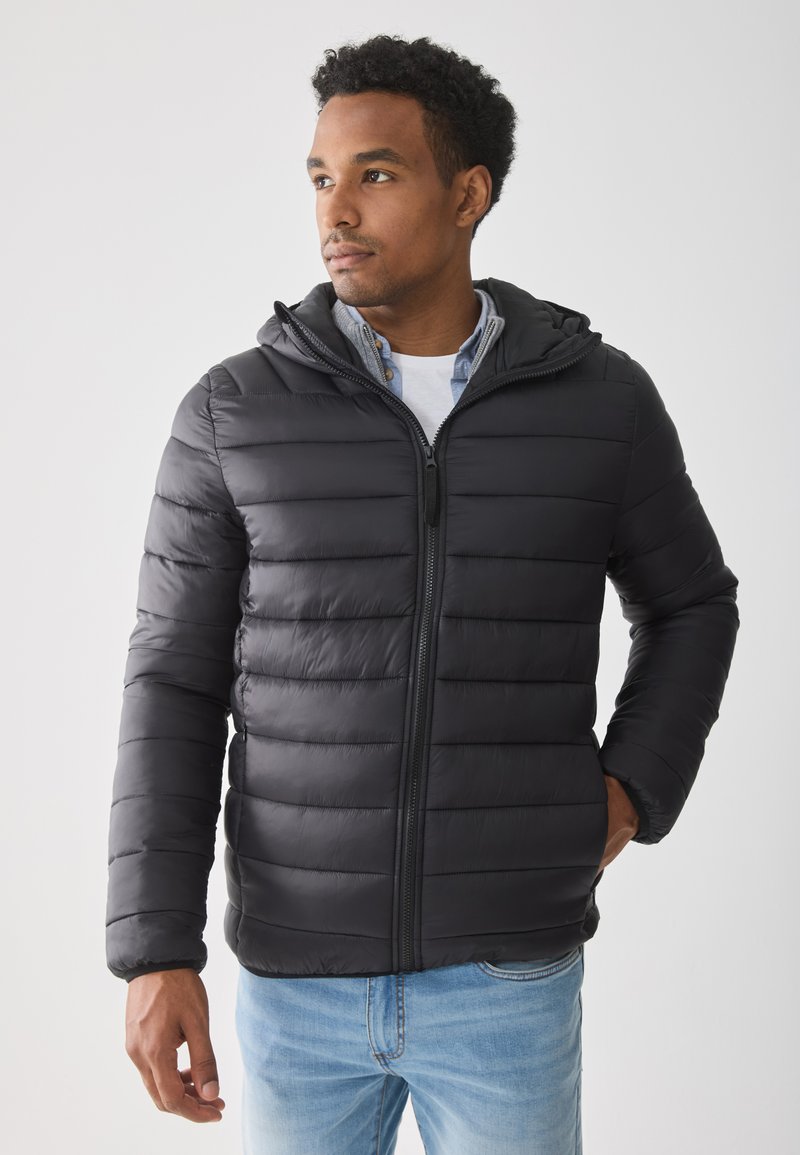 Hombre con una chaqueta acolchada negra con capucha sobre una camisa azul y una camiseta blanca, de pie con una mano en el bolsillo, mirando a la izquierda.
