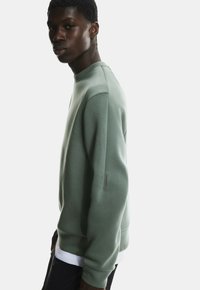 Zielony sweter o teksturowym wykończeniu, z okrągłym dekoltem i subtelnym szwem na rękawie. Nosi się go na białej koszuli.