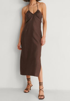 Kobieta ubrana w bezrękawnikową, ciemnobrązową sukienkę midi na ramiączkach z marszczonym dekoltem, w brązowe sandały na wysokim obcasie na rzemykach, stojąca na tle białej ściany.