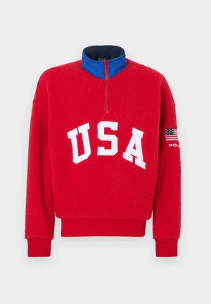 Červený fleecový pulovr s polovičním zipem na límci, bílým nápisem "USA" a modrým vnitřním límcem. Má nášivku s vlajkou na rukávu a žebrované manžety.