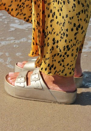 Sandales à plateforme dorées avec deux boucles, assorties à une jupe plissée jaune avec des taches noires, debout sur une plage de sable près de l'eau.