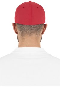 Piros baseball sapka strukturált koronával és lapos karimával, visszafordítva viselve. Fehér poloing látható a nyaknál, rövid haj alatta.