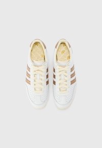 Adidași albi cu accente din piele maro și trei dungi, având un model perforat, șireturi crem și o branț galben cu logo.