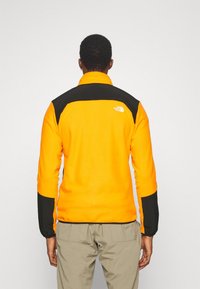 Giacca in fleece di un vivace arancione con dettagli neri, caratterizzata da un alto colletto e polsini a costine. Logo visibile sulla parte superiore della schiena.