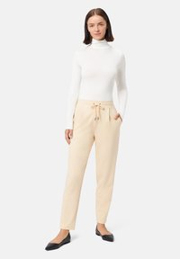 Pantalones beige con cordón, bolsillos laterales y piernas entalladas, combinados con un suéter de cuello alto ajustado blanco y zapatos planos negros.