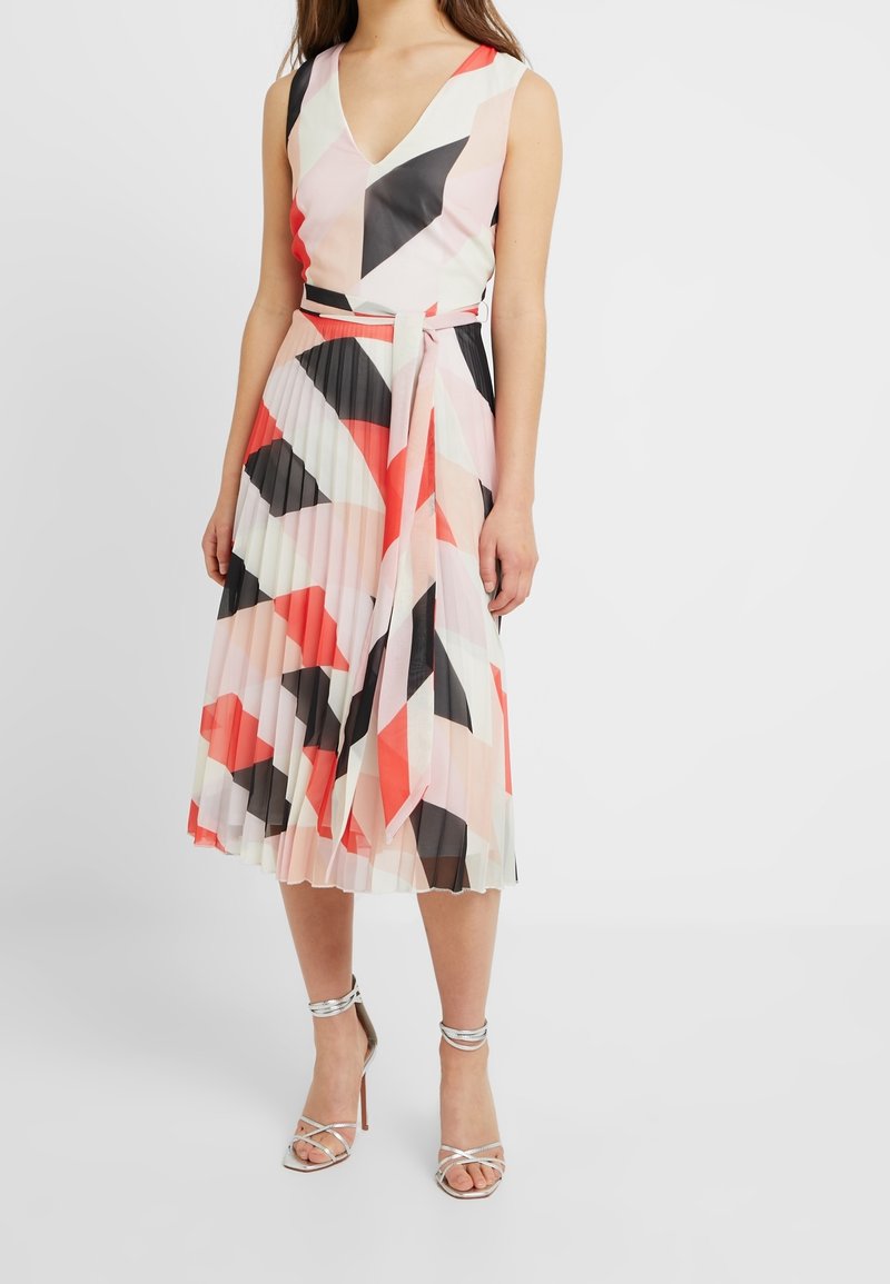 Mujer vistiendo un vestido midi sin mangas, con pliegues y un patrón geométrico en coral, negro, blanco y rosa, combinado con tacones de tiras plateadas en el tobillo.