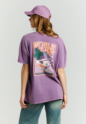 Femme portant une casquette violette et une chemise violette oversize avec un trèfle vert et le texte "Que la chance soit de votre côté" au dos, associée à un jean bleu.