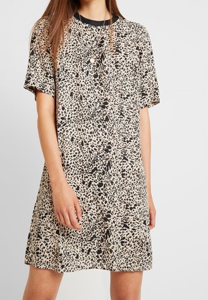 Kvinde iført beige og sort leopardprint kortærmet kjole med rund halsudskæring og lagdelte guldkæder, stående foran en ensfarvet baggrund.