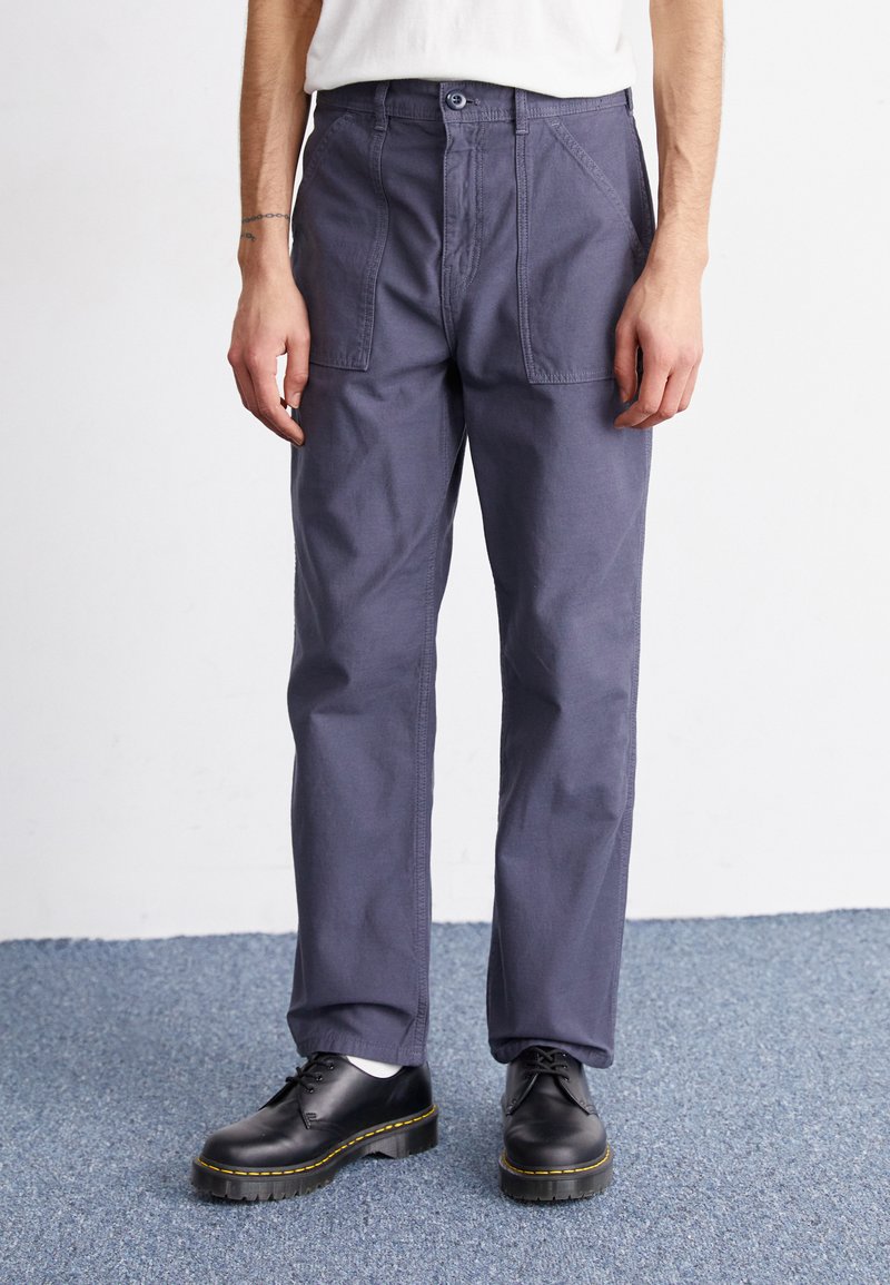 Navy blue trousers in a relaxed fit, made from lightweight fabric. Features front pockets and a button closure. Paired with black lace-up shoes.
